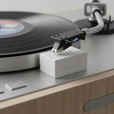 Record player stylus gently touching a small white piece of Mr. Clean Magic Eraser on a turntable platter, illustrating a common DIY cleaning method with soft, even lighting.